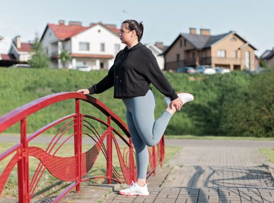 A woman in active wear performs a stretching exercise on a red bridge in a suburban park setting.