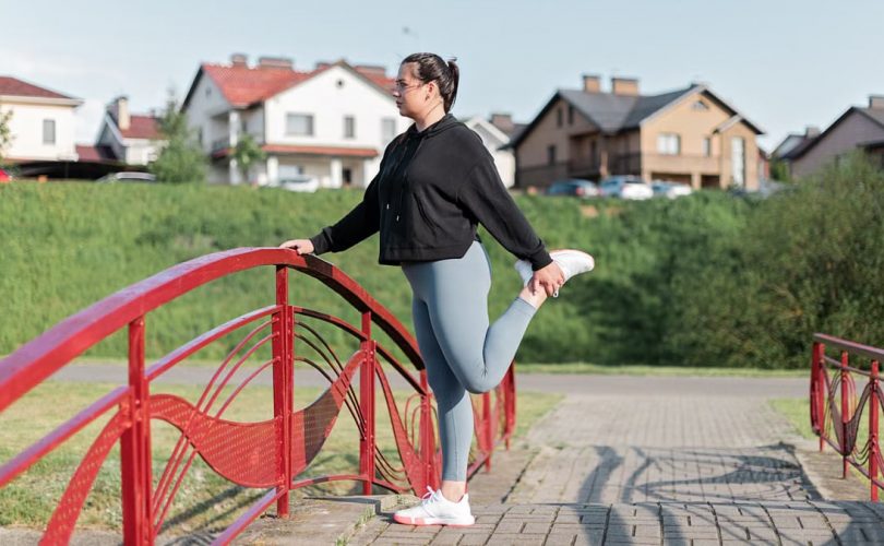 A woman in active wear performs a stretching exercise on a red bridge in a suburban park setting.